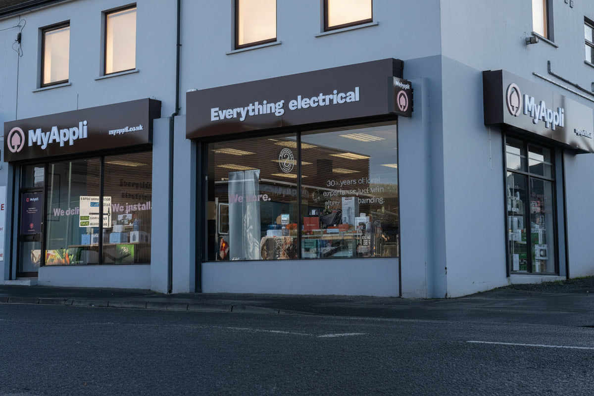 A white two-story commercial building with storefronts, including a sign reading 'MyAppli' and another for 'Everything electrical', set against a pale sky with wet pavement in the foreground.